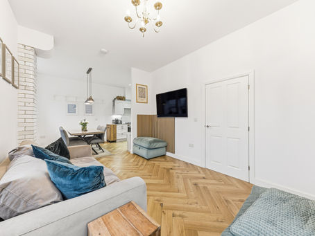 Modern living room with gray sofa, teal pillows, and a TV on the wall. Wooden herringbone floor, dining area, and kitchen in the background.