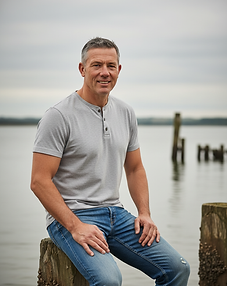a_rugged_outdoor_portrait_of_a_man_sitting_on_a_weathered_wooden_post_near_the_water_he_we