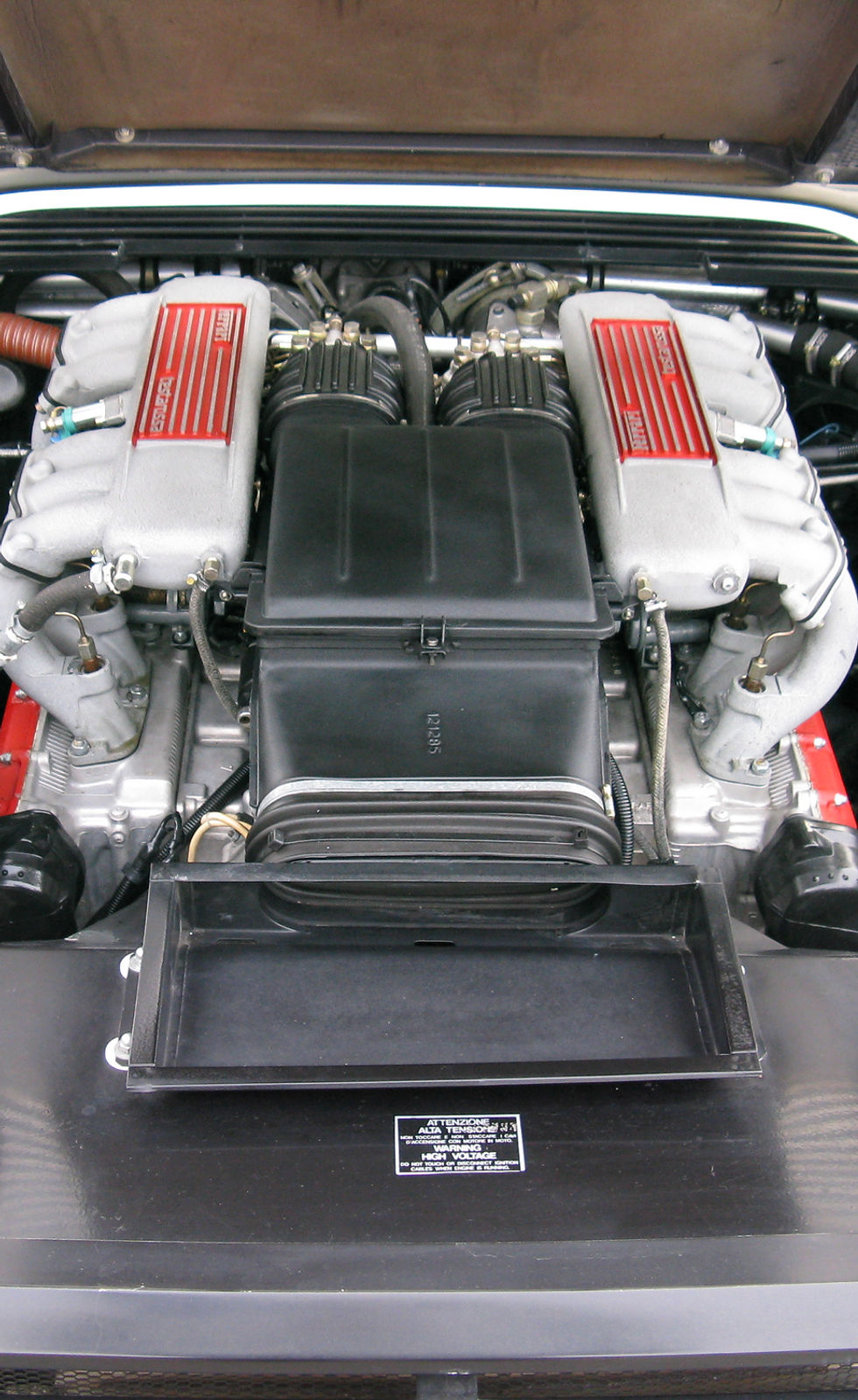 Top-down view of a classic sports car engine bay with many mechanical details, mostly black and metallic with some red accents, landscape orientation.