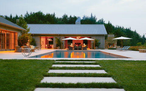 A concrete path in the grass that leads to a large swimming pool with umbrellas and outdoor lounge seating around