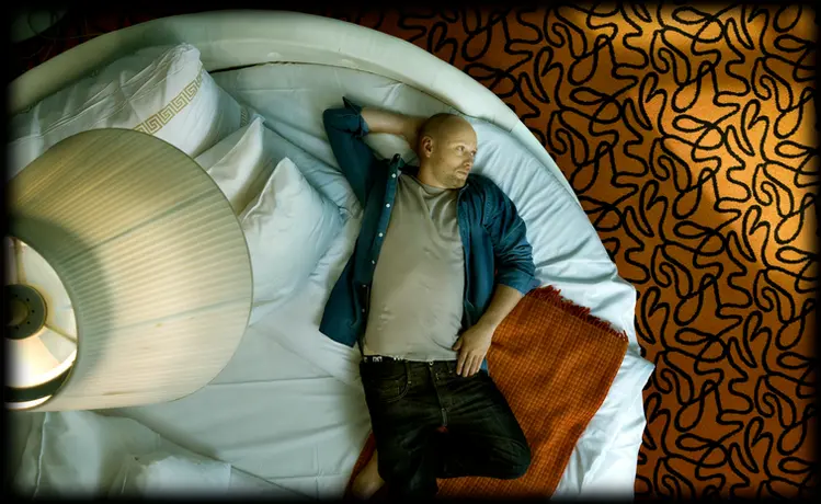 An environmental portrait of Norwegian actor Axel Hennie, taken from above as he lies on a large white round bed in a hotel suite in Oslo. A lamp is positioned above him, and an orange carpet serves as the background.