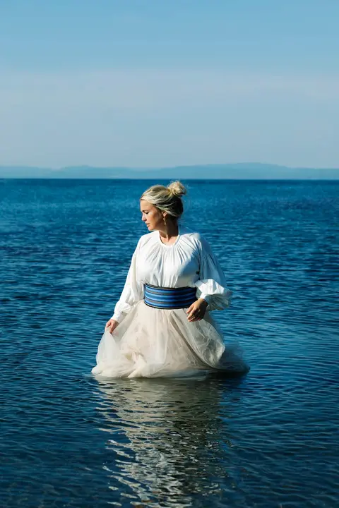 A portrait of Norwegian singer Helene Bøksle, dressed in a stylish outfit, with her dress getting wet as she stands in the ocean, waves gently surrounding her.