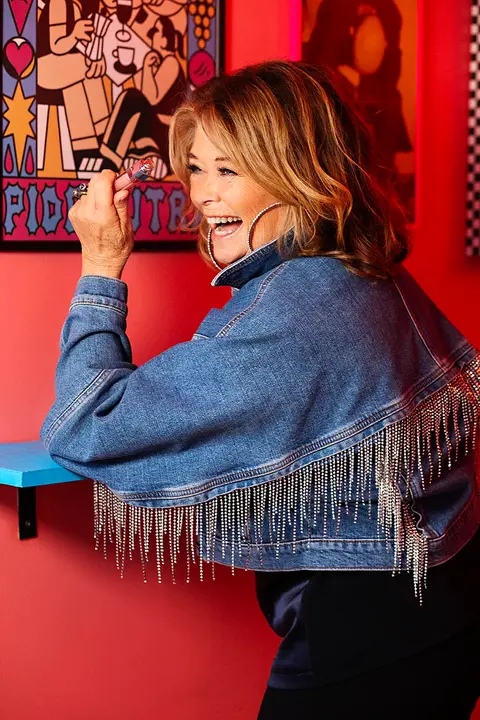 Close-up of pop artist Wenche Myhre leaning on a blue bar table, laughing in a colorful 80s-inspired venue.