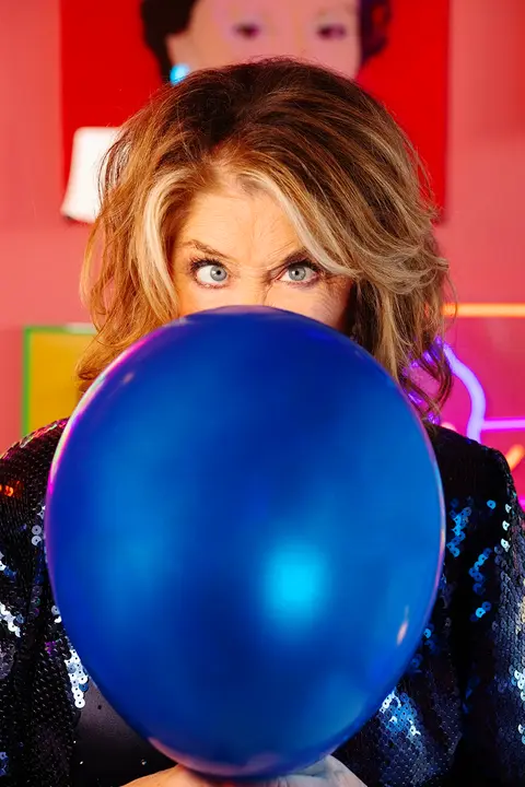 Portrait of Norwegian pop star Wenche Myhre, behind a blue balloon in a colorful 80s-inspired venue.