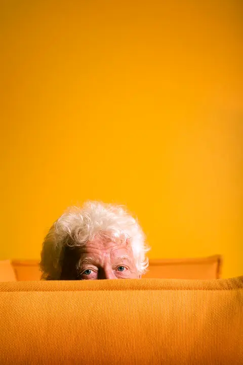 A portrait of Norwegian journalist Odd Karsten Tveit, with half of his face visible behind a bright yellow sofa, set against a yellow backdrop.