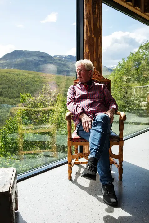 Portrait of chef Arne Brimi sitting in a chair at his restaurant, Vianvang, with large windows behind him overlooking a scenic landscape. He is gazing thoughtfully out the window.