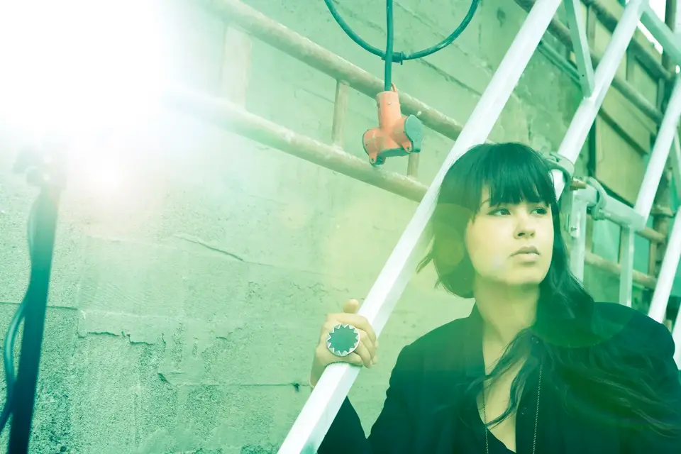 Pop singer Maria Mena, sitting on a stair against a brick wall, holding the rail and looking away from the camera.