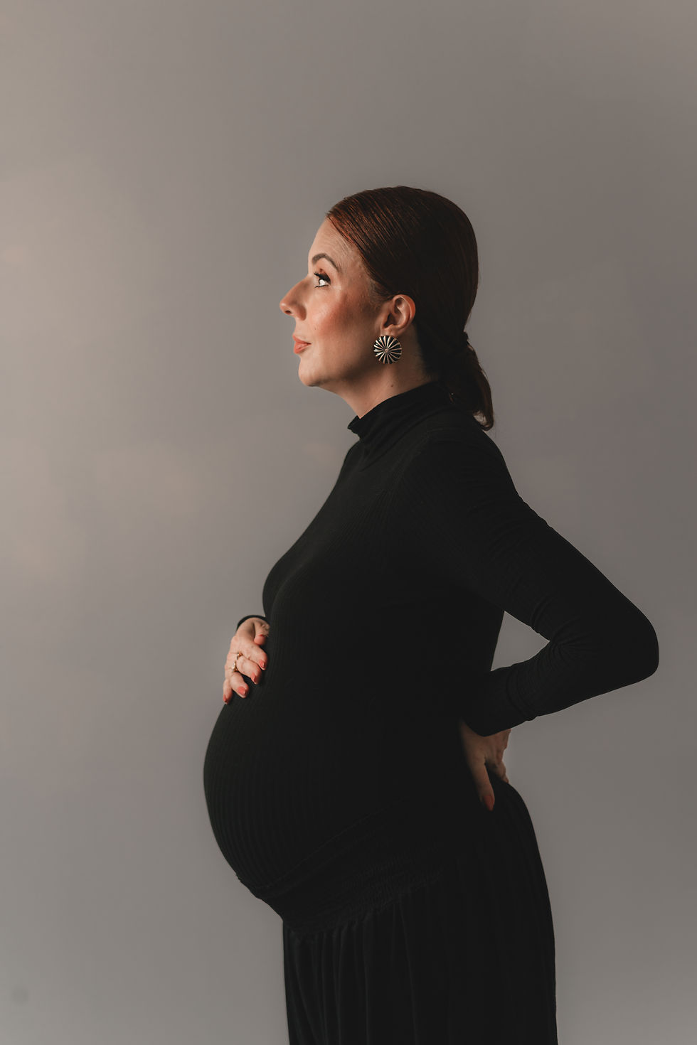 Grávida de perfil, de vestido preto, posa com as mãos na barriga em estúdio cinza, com expressão serena. crônicas de maternidade depois dos 40