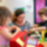 Woman supervises three young children playing with toy cash register and food.