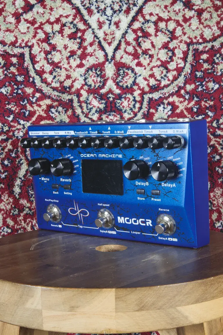 Mooer Ocean Machine pedal on a wooden stool with patterned rug backdrop, angled to the left.