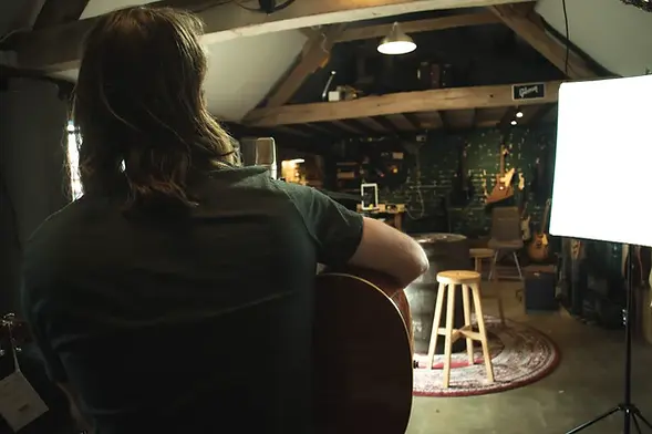 A songwriter looking out at the rustic interior of a guitar shop while recording their music.