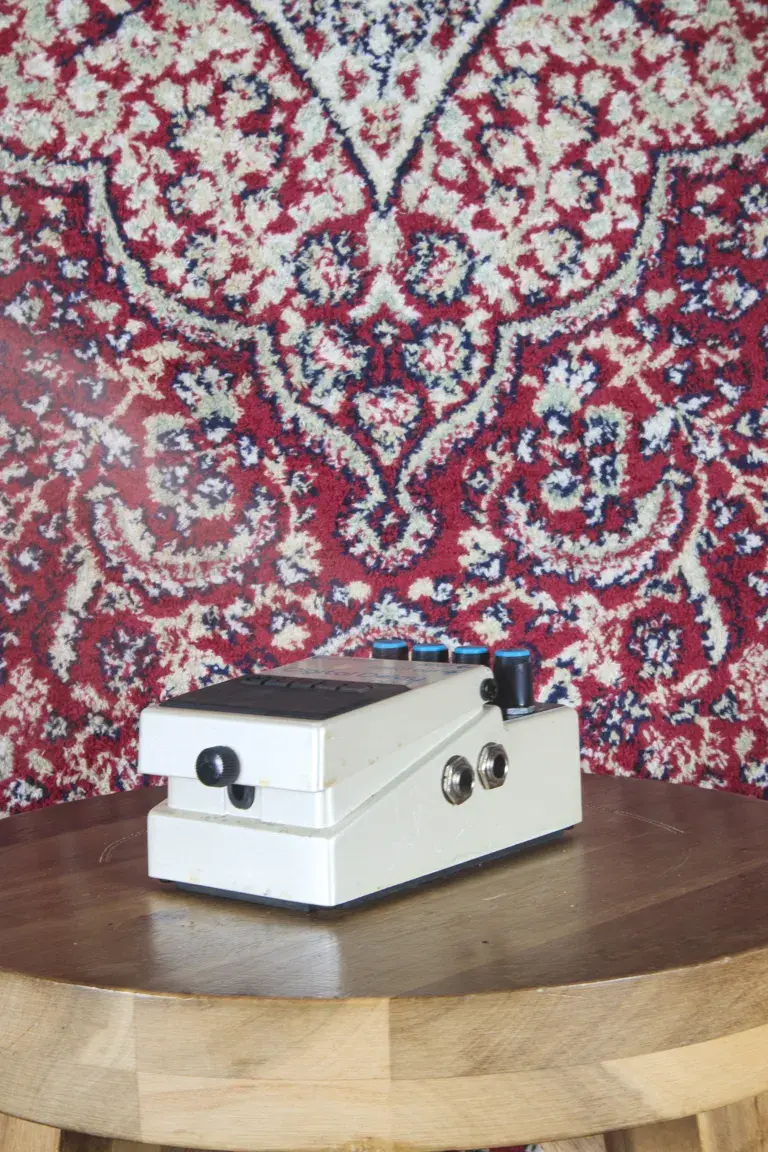 Boss DD-3 Digital Delay pedal on a wooden stool with patterned rug backdrop, angled to the right.