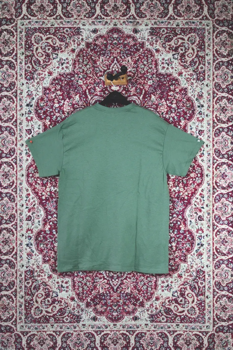 Back view of sage green Americana tee laid flat on red patterned rug