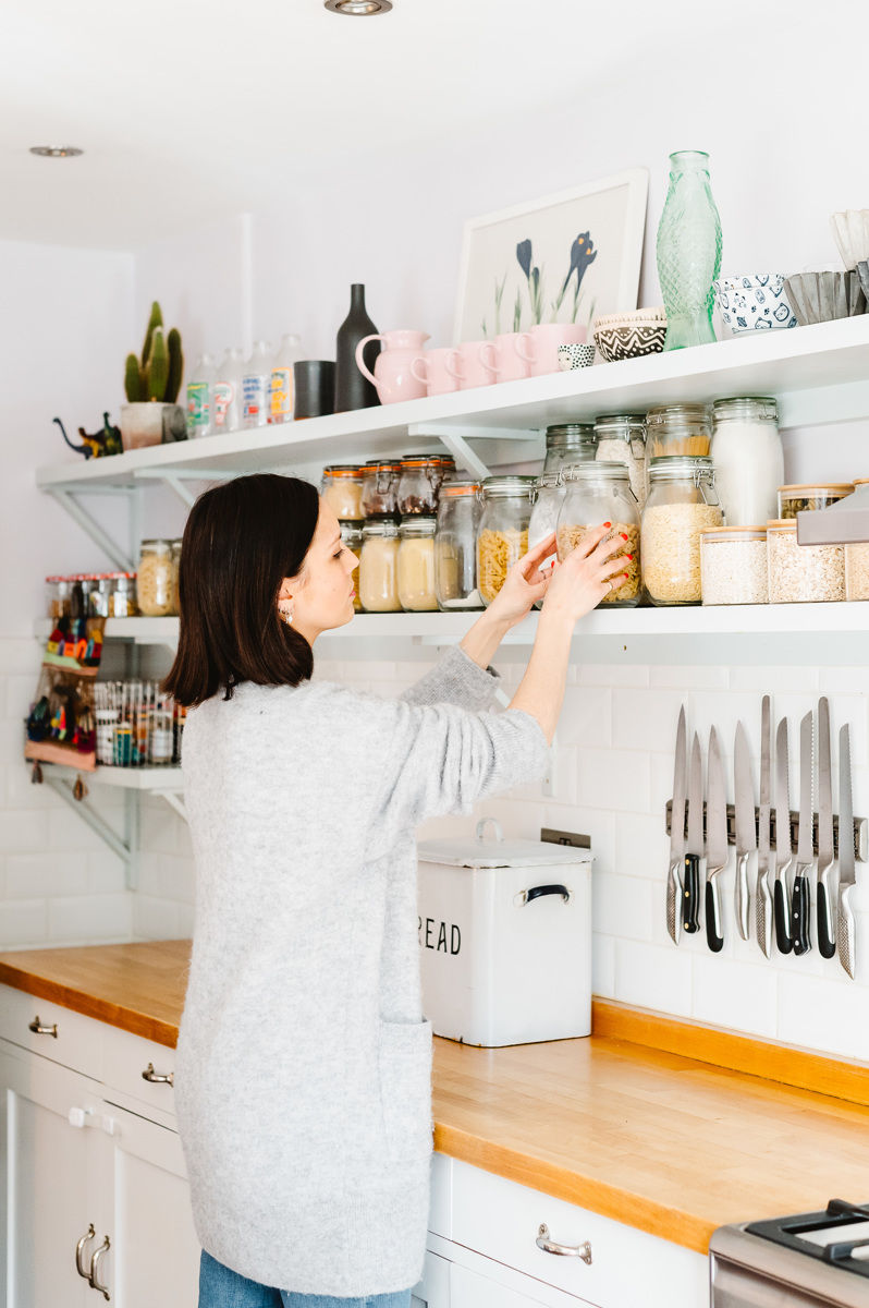 Woman in gray sweater organising glass jars on a kitchen shelf. White cabinets, knives on wall. Bright, tidy atmosphere.