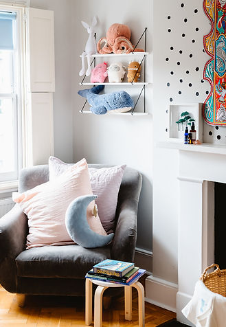 The corner of a child's bedroom, the white walls have 3 white shelves displaying well arranged soft toys. There is a grey armchair in the corner with 3 big chshions and a blue moon shaped cushion on it and books arranged nicely on a wooden stool next to the chair