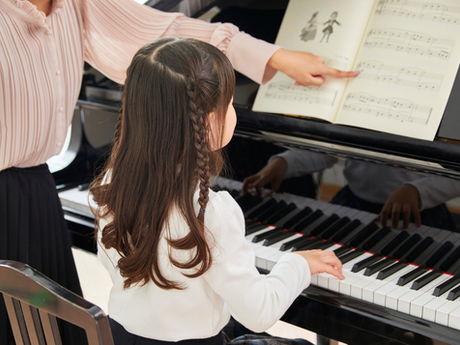A young girl during her piano lesson, her teacher is pointing at the page while she follows along and play.