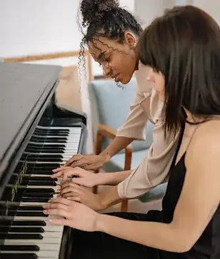 An adult piano student with her hands on the piano keys as guided by her piano teacher