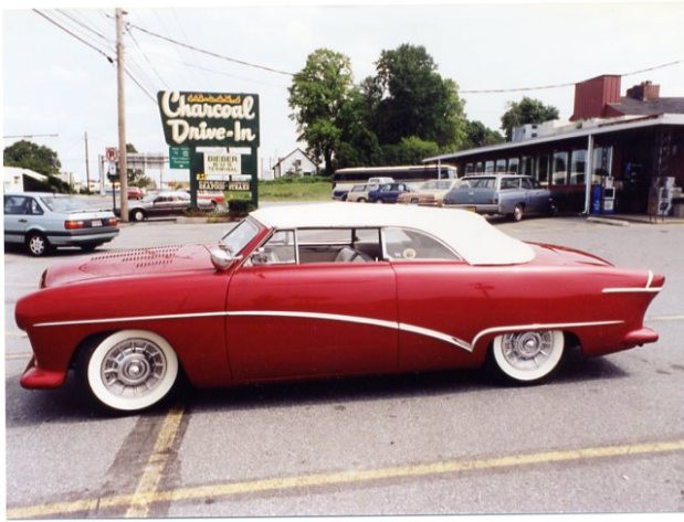 a bright red car sits in the parking lot of a restaurant called charcoal drive-in. other cars are parked behind it in the distance, but the focus is on the custom ford in the foreground. the image is taken of the side to show the full shape of the body, the detailing, the whitewashed wheels, and the convertible top