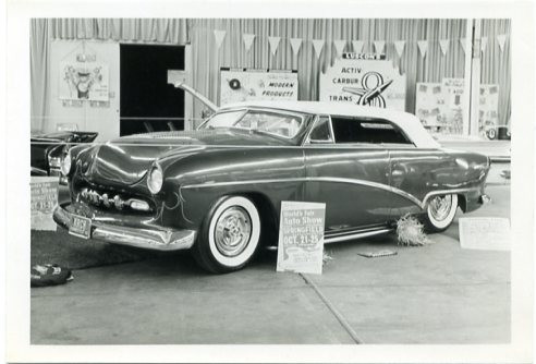 a black and white photo of a custom Ford. it is on a showroom floor with a sign (unreadable) by the front wheel. the background has many signs (unreadable) and flag garlands hanging on a wall that seems to have a curtain covering it