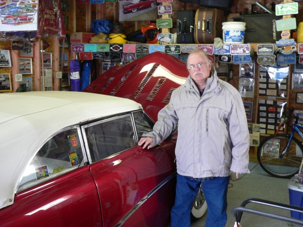 ray soff, an older man with a handlebar mustache wearing a coat, jeans, and glasses, touches the red ford by the front passenger side window. behind him is a cluttered but organized wall of items related to cars like license plates and photos