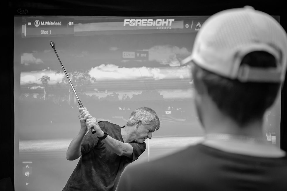A golfer at the top of a backswing hitting a ball into a simulator screen while being watched by a golf instructor.