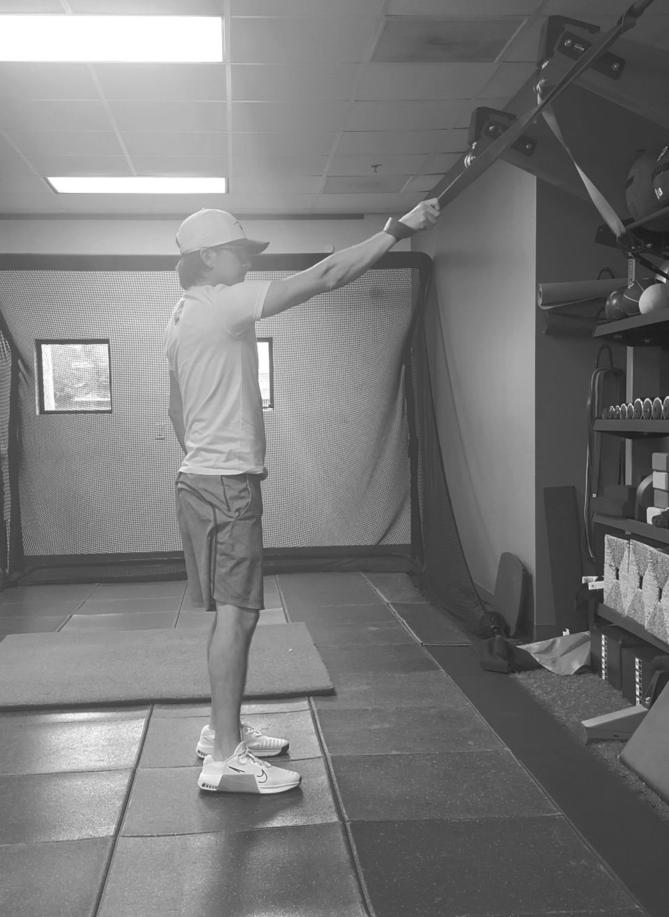 A man in a white shirt and red shorts is about to stretch with a band. There’s a golf net and windows in the background.