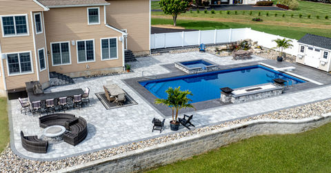 Aerial view of a custom backyard in Halfmoon, NY with in-ground pool, raised spa, paver patio, outdoor bar, fire pit seating, and retaining wall behind a two-story home.