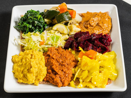 A square plate with colorful Ethiopian dishes like lentils, greens, beets, and cabbage on injera. Bright, vibrant colors on a dark background.