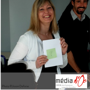 Photo de Joson Deforas : Une personne très souriante au premier plan montre une feuille sur laquelle est affichée un post-it avec un point d’interrogation. Une seconde personne derrière regarde l’objectif en souriant.