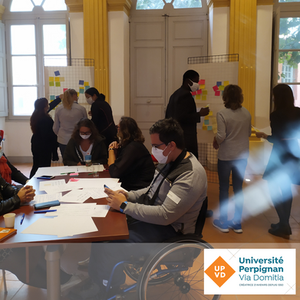 Atelier participatif. AU premier plan plusieurs personnes autour d'une table. Au deuxième plan deux petits groupes étudient des paper board avec des post-its.
