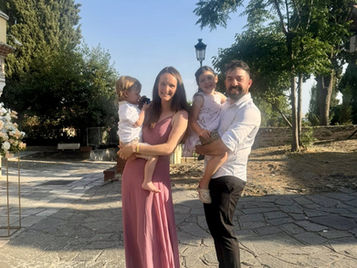 A photo of a family of four dressed in summery formalwear at a Greek wedding. The parents are smiling, holding their two young children, with olive trees and whitewashed buildings in the background, capturing the warmth and joy of the celebration.