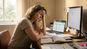 Person sitting at a desk with spreadsheets, a laptop, and dual monitors while resting their head in one hand.