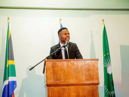 Photograph of Reinhold Mangundu speaking at a formal podium, flanked by the Namibian and African Union flags.