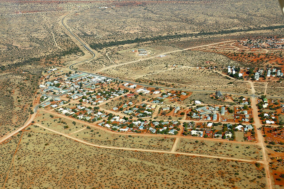 Wide high-angle aerial photograph of the town of Aranos in Namibia, showing residential streets, small buildings, and dirt roads surrounded by the expansive, dry, orange-red sand dunes and sparse bush terrain of the Kalahari desert.