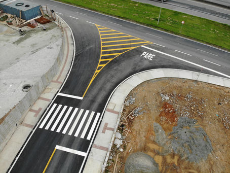 Imagem aérea de um acesso viário recém-implantado, com pavimento asfáltico novo e sinalização horizontal destacada. Há faixa de pedestres branca, marcação de “PARE” no solo e área zebrada amarela canalizando o fluxo de veículos em uma conversão. O entorno apresenta canteiros e áreas em obra, com trechos de terra exposta e infraestrutura viária ao fundo.