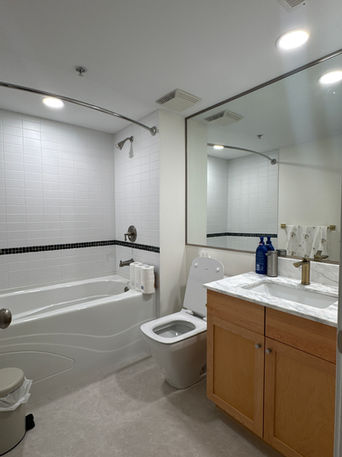 A clean, minimalist bathroom with white tiled walls, a bathtub and shower combo, a marble countertop vanity, and warm wood cabinetry.