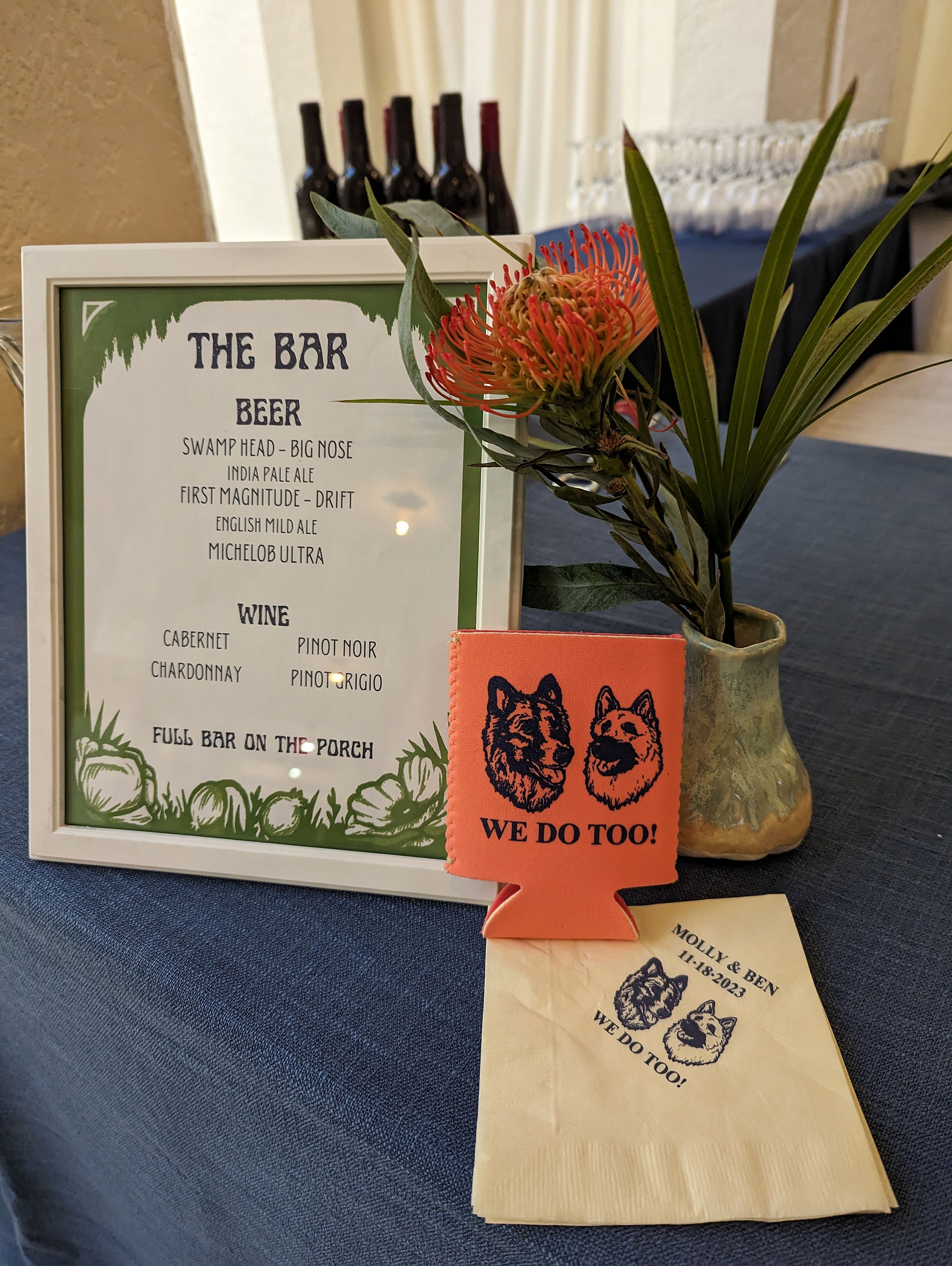 A frame a list of signature cocktails on it with a bouquet of flowers, a napkin, and a koozie next to it