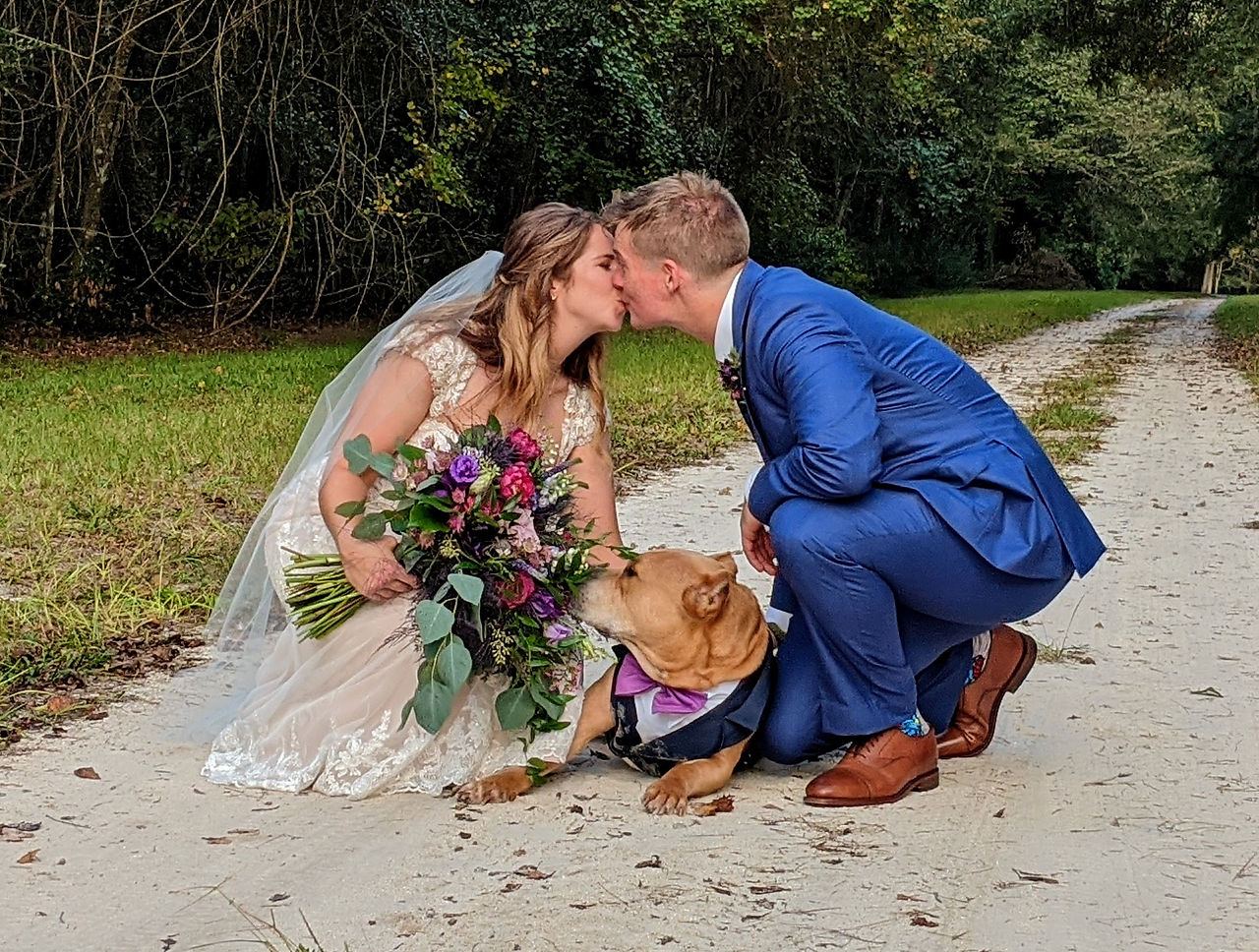 A bride and groom crouched down by their dog while kissing
