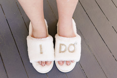 A bride's feet in white fuzzy slippers that say "I Do"