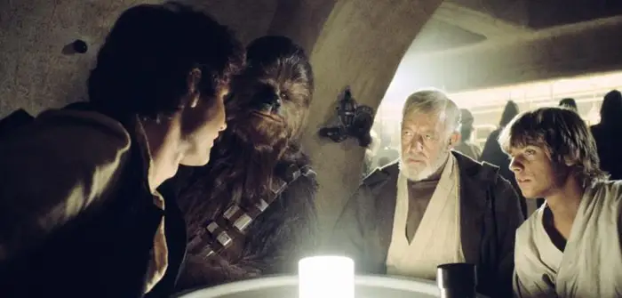 Luke, Chewbacca, Obi-Wan and Han sit tense around a glowing table in a dim cantina, with onlookers in the background.