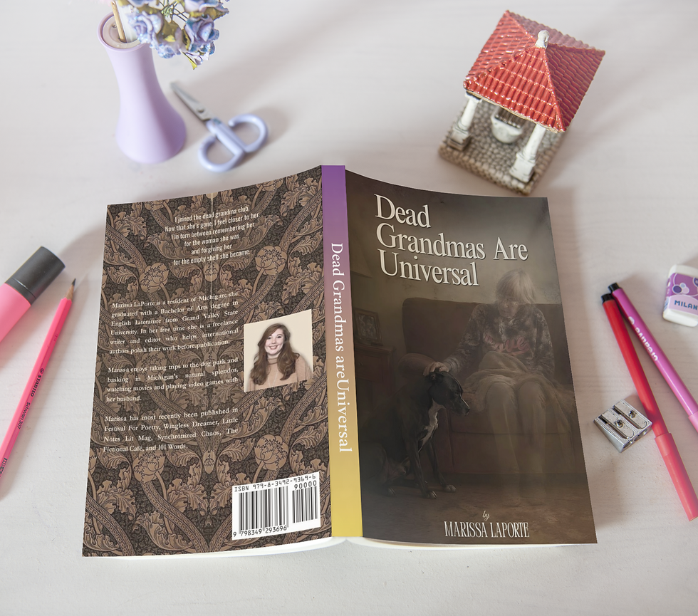 Open book titled Dead Grandmas Are Universal on a white desk, surrounded by pencils, scissors, a vase, and a small house model.