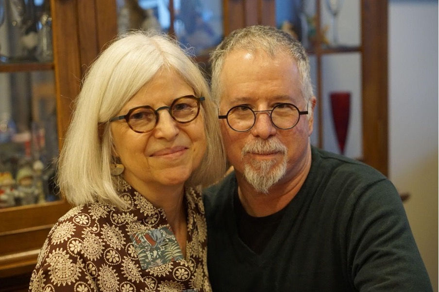 Smiling older couple with glasses pose indoors by wooden cabinets, warm light and cozy mood.