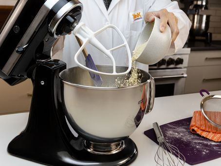 Person in a Think Ingredients lab coat pours dry ingredients into a stand mixer in a kitchen.
