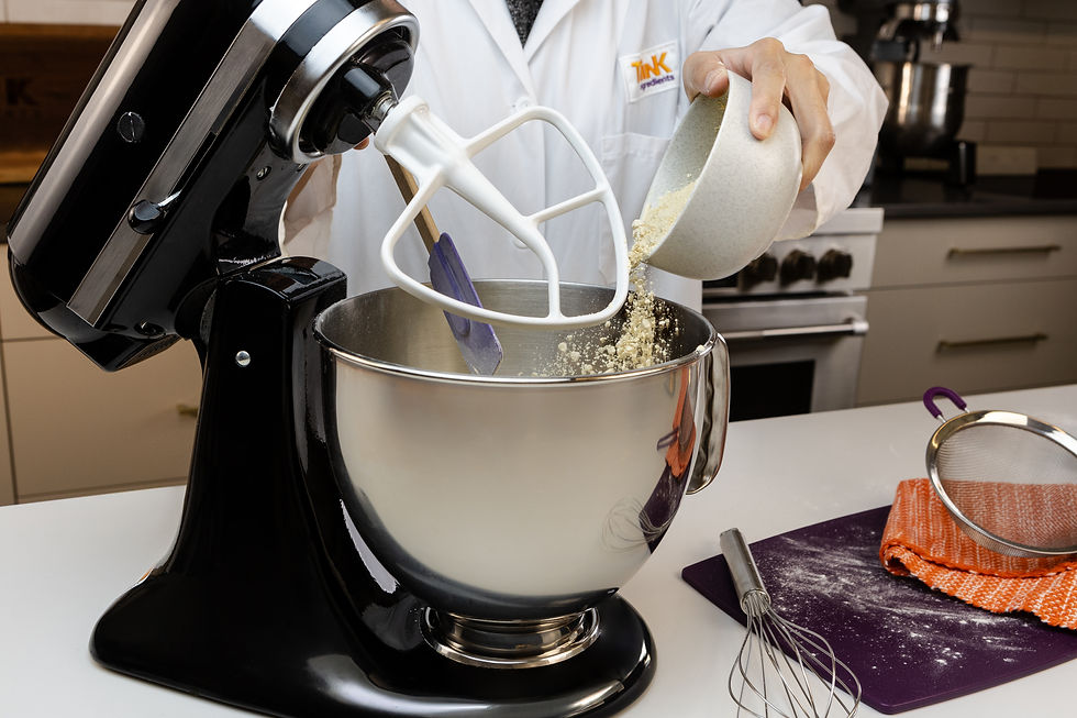 Person in a Think Ingredients lab coat pours dry ingredients into a stand mixer in a kitchen.
