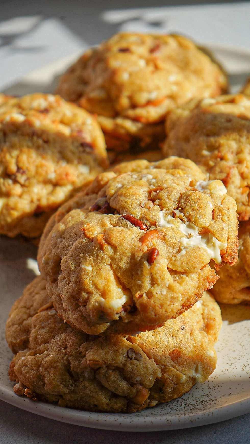 Close-up of chocolate chip cookies with yellow and brown chips on a white plate. Blurred brown cup and white bag in the background.