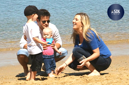 Blonde woman in blue shirt crouching on beach in front of man with dark hair and sunglasses, small infant in pink shirt, and toddler wearing black hat