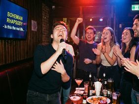 Junger Mann singt mit Mikrofon „Bohemian Rhapsody", Freund:innen jubeln und applaudieren am Tisch mit Cocktails – Karaoke-Abend in Wien