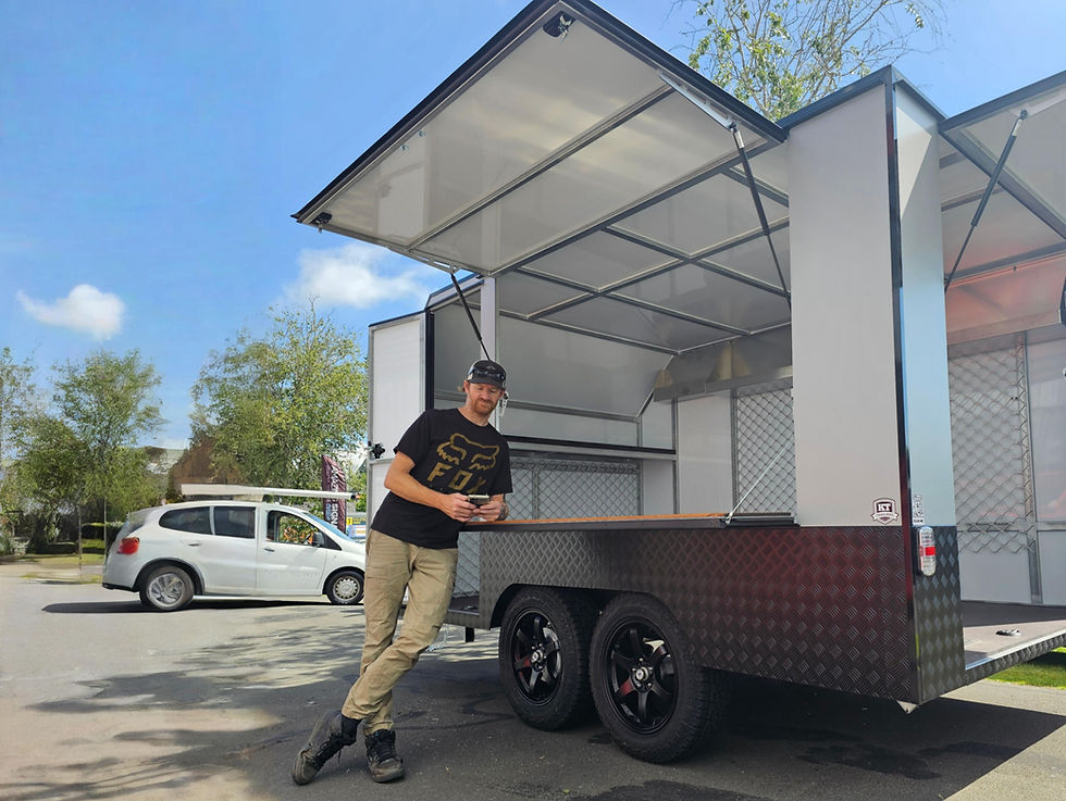 Man leaning on a grey trailer with open doors. Parked white car and trees in the sunny background.