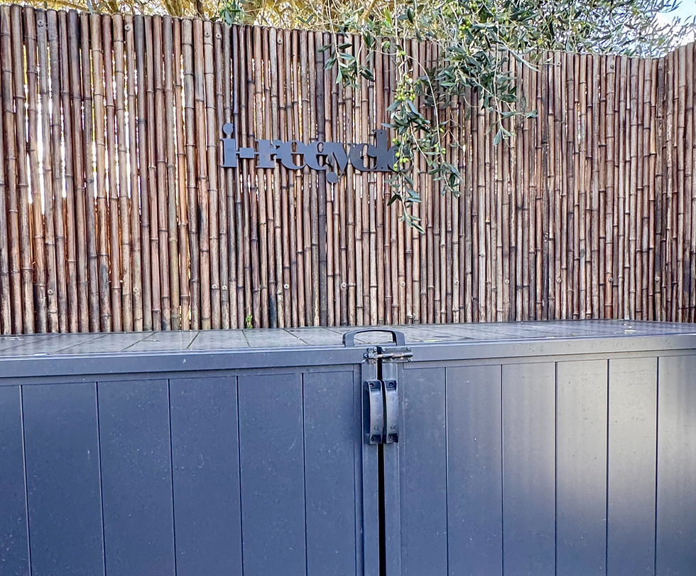Black storage box in front of a bamboo fence with "i-recycle" written on it. Leaves hang overhead in a sunny outdoor setting.