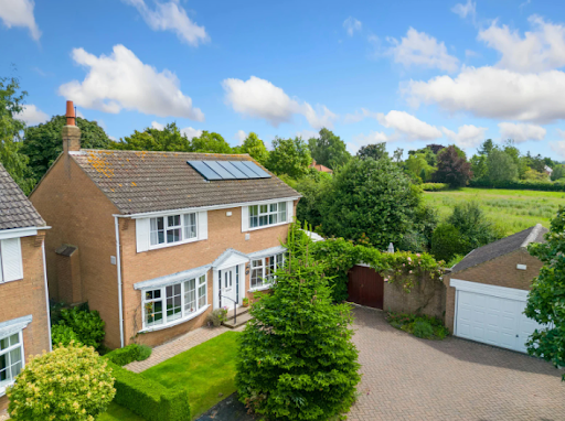 Detached brick home with rooftop solar panels, driveway and garage, surrounded by green lawn and trees under a bright blue sky.